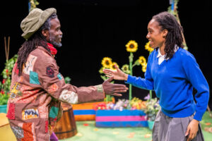 Two Black people on a stage set with bright flowers and sunflowers, reaching out to high-five. The person on the left wears a brown patched jacket, a red scarf and a green bucket hat. The person on the right wears a blue V-neck sweater over a white shirt and a grey skirt, smiling widely.