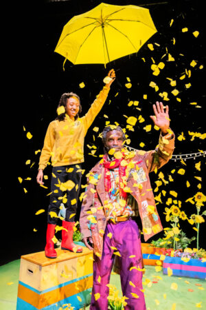 A stage performance scene featuring two Black people in a shower of yellow flower petals. The person on the left stands on a colourful box, wearing a yellow hoodie and red Wellington boots, holding a yellow umbrella high above the other person. The person in front is wearing a brown patched jacket and purple trousers, with one hand raised and looks directly at the camera.