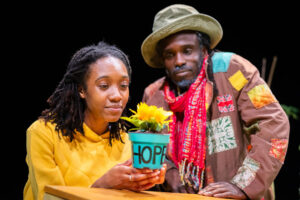 A close-up of two Black people looking at a small teal flowerpot with a yellow flower, which has the word 