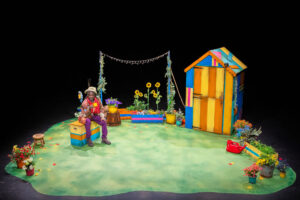 A wide shot of a brightly coloured stage set featuring a sunny garden scene against a black backdrop. The set includes a tall, multi-striped shed, sunflowers, planters, a wooden barrel and string lights. A Black person wearing a brown patched jacket, purple trousers, a red scarf and a hat is seated on a bright yellow and blue box on the left side of the stage.
