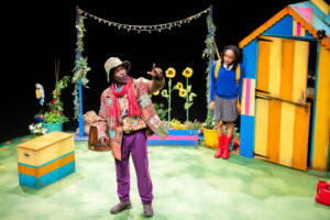 A Black person in a brown patched jacket, purple trousers, a red scarf and a light-coloured bucket hat stands in the centre of the stage, gesturing with a pointing hand. Behind them to the right, another Black person wearing a blue sweater, grey skirt, red Wellington boots and a backpack stands looking over their shoulder toward the foreground. A brightly striped shed and sunflowers are visible on the stage set.
