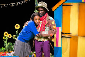 Two Black people on stage embracing. The person on the right wears a brown patched jacket, purple trousers, a red scarf and a bucket hat, smiling warmly. The person on the left wears a blue sweater and grey skirt, clinging to the other person's torso. A brightly striped shed and sunflowers are in the background.