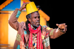 A close-up of a Black person on stage wearing a brown patched jacket, a red fringed scarf and holding a yellow bucket on top of their head like a hat. They have a surprised or dramatic expression and are extending one hand out, mid-performance.