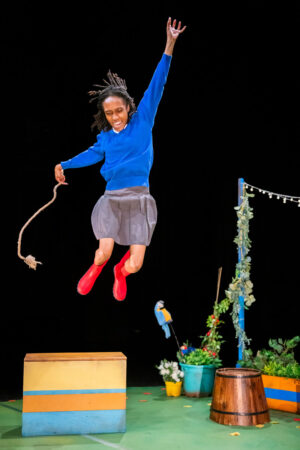 A Black person in a blue long-sleeved shirt, grey skirt and red Wellington boots jumps high in the air, with their right hand extended upwards and a rope dangling from their left. Below them is a brightly coloured box and a wooden barrel, part of a cheerful garden-themed set against a black backdrop.