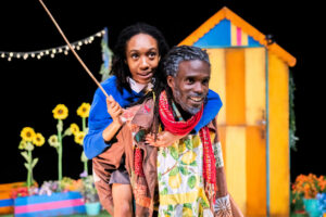 A close-up of two Black people having a piggyback ride on a colourful stage set with sunflowers and a striped shed. The person carrying wears a brown patched jacket with a lemon-print panel and a red scarf, smiling widely. The person on their back wears a blue sweater and a brown skirt, holding onto a thin stick.