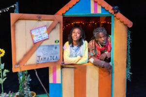 Two Black people look out from the open door of a brightly coloured, striped shed on stage. The open door reveals a sign with a map of Jamaica and the word 