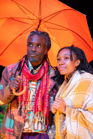 A close-up of two Black people sharing a bright orange umbrella. The person on the left, wearing a brown patched jacket and a fringed red scarf, holds the umbrella handle and looks up. The person on the right is wrapped in a plaid blanket and looks to the side with a serene expression.