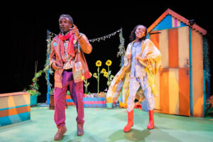 Two Black people are standing on a brightly lit stage with a garden set, including a striped shed and sunflowers. The person on the left wears a brown patched jacket, purple trousers and a red scarf, looking up with an open mouth. The person on the right wears patterned pyjamas, red rubber boots and a plaid blanket draped over their shoulders, with a wide, joyful expression.
