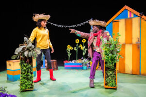 Two Black people on stage in a bright garden setting with a shed and sunflowers. Both wear straw hats. The person on the left wears a yellow hoodie, black trousers and red Wellington boots. The person on the right wears a patched jacket, purple trousers and a red scarf, dramatically pointing to the right. Both stand next to tall planters wrapped in greenery.