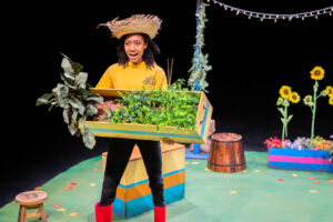 A Black person on stage wearing a straw hat, a yellow hoodie, black trousers and red Wellington boots. They are holding a large, wooden planter filled with vibrant green plants and looking forward with a surprised expression. The stage set features a green floor, a multicoloured box, a small stool and sunflowers in the background.