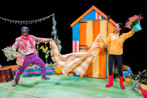 Two Black people on stage holding a large, yellow and white plaid blanket spread out between them, standing in front of a multicoloured shed. The person on the left wears a brown patched jacket and purple trousers, holding a bunch of flowers. The person on the right wears a yellow hoodie and red Wellington boots, holding a potted pink flower up high.